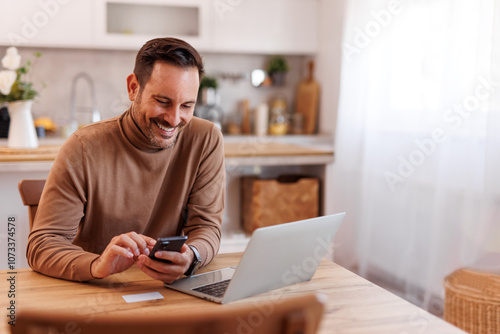 Portrait of smiling mid adult man with credit card using mobile phone and laptop on dining table at home