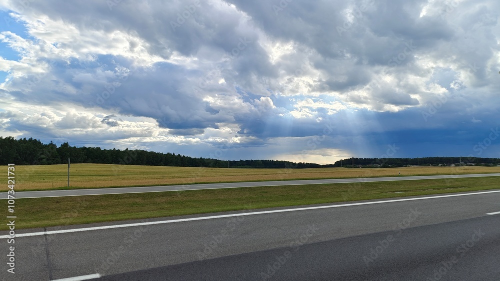 Fototapeta premium An asphalt road with markings is laid out among agricultural fields. A power line runs nearby, and behind it a forest grows and a village stands. On a summer day, sunlight breaks through thick clouds