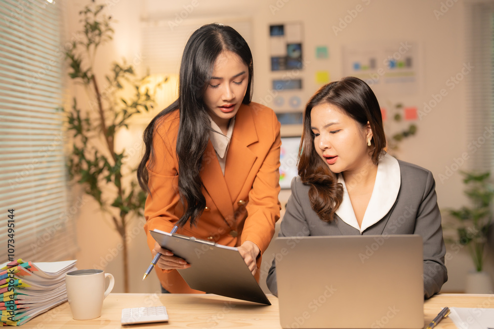 Two asian businesswomen are discussing work, with one pointing at a document on a clipboard and the other looking at a laptop, in a modern office environment Two asian businesswomen are discussing work, with one pointing at a document on a clipboard and the other looking at a laptop, in a modern office environment