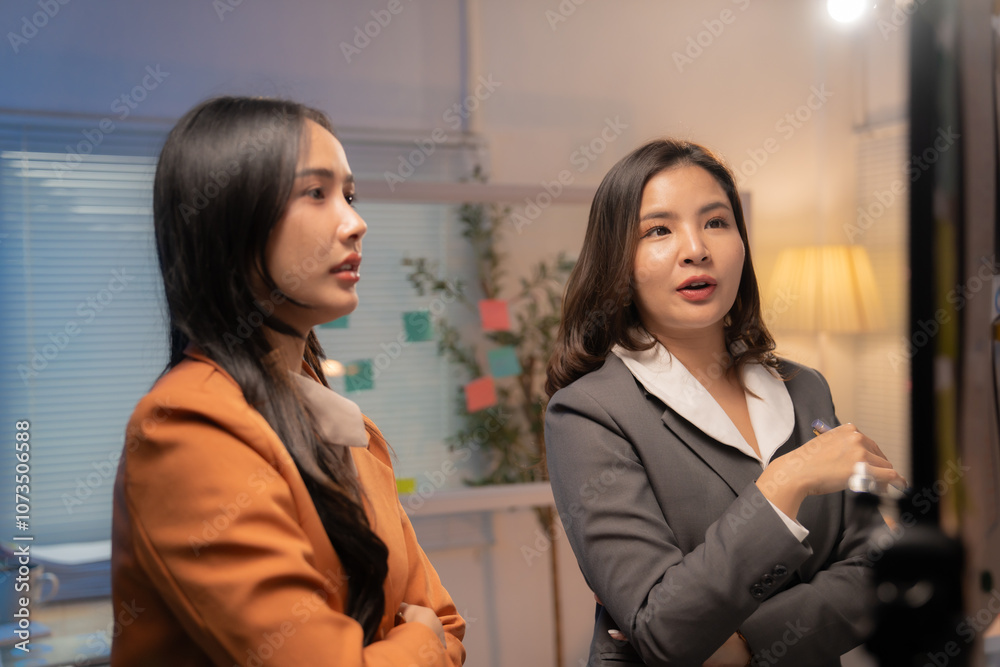 Two asian businesswomen wearing suits are standing in an office, actively engaged in a video conference, discussing a project with gestures and focused expressions Two asian businesswomen wearing suits are standing in an office, actively engaged in a video conference, discussing a project with gestures and focused expressions