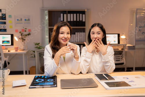 Two asian businesswomen discussing positive financial results in office at night, one explaining while the other is surprised, covering her mouth