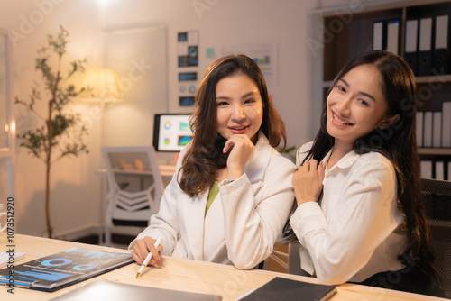 Two young businesswomen are sitting at a desk, working late in a brightly lit office, with documents and a computer visible