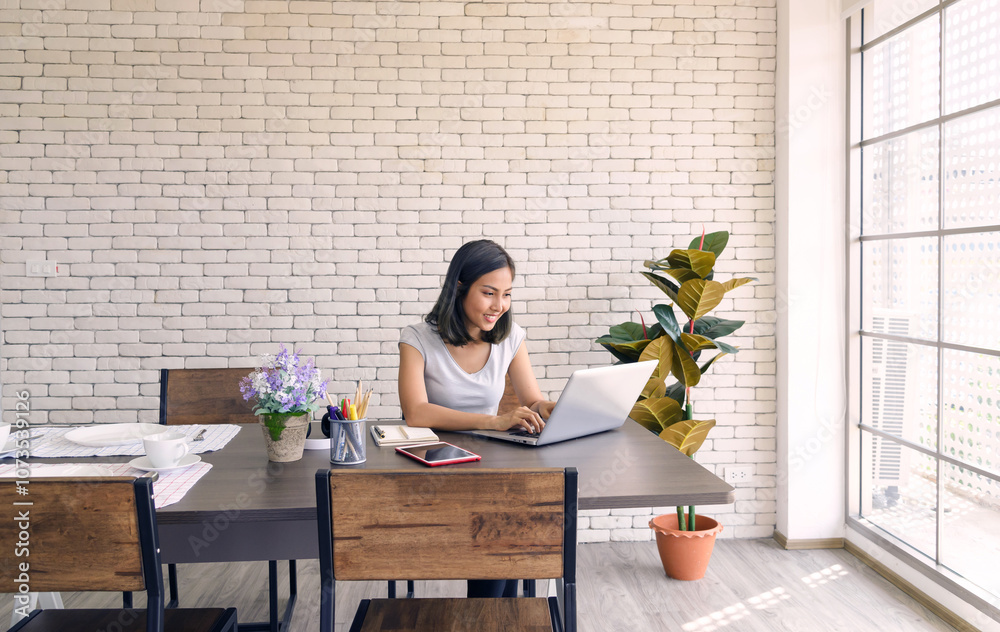 Young asian woman in casual clothing sitting at big wooden table in modern kitchen, working with computer laptop at home. Work at home concept.
