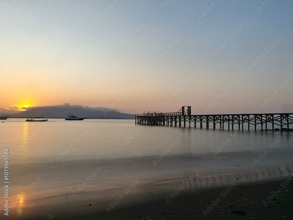 A rustic wooden pier stretches into the calm waters of a bay at sunrise, with a boat anchored in the distance and distant mountains silhouetted against the sky. A rustic wooden pier stretches into the calm waters of a bay at sunrise, with a boat anchored in the distance and distant mountains silhouetted against the sky.