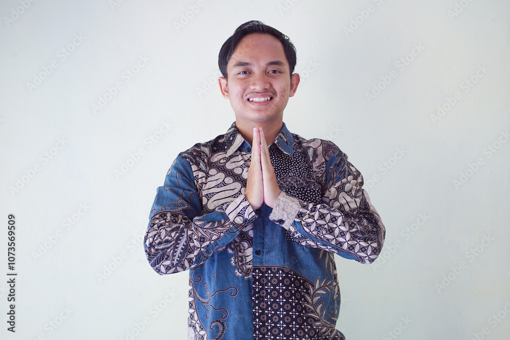 Smiling Asian man wearing batik shirt, showing hand gesture of apology and welcome. Advertising concept. Smiling Asian man wearing batik shirt, showing hand gesture of apology and welcome. Advertising concept.