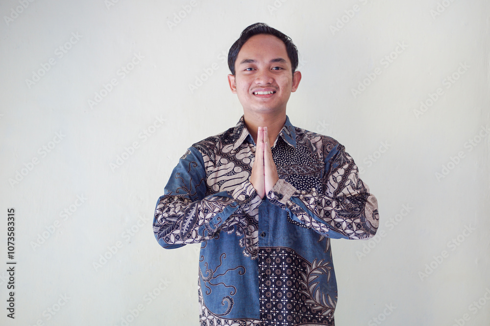 Smiling young teacher in batik shirt with hands as traditional greeting gesture, expressing warmth and friendliness, standing against plain background. Smiling young teacher in batik shirt with hands as traditional greeting gesture, expressing warmth and friendliness, standing against plain background.