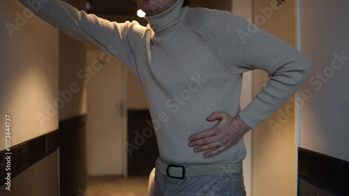 A person in a beige turtleneck and plaid trousers is standing in a hallway, holding their lower back as if experiencing discomfort. The warm lighting adds an introspective, moody atmosphere.





