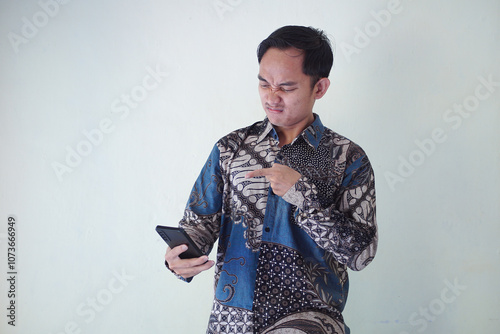 Shocked Asian man wearing batik shirt and looking at his cellphone screen, isolated on white background. expressing surprise and curiosity in a relaxed atmosphere.