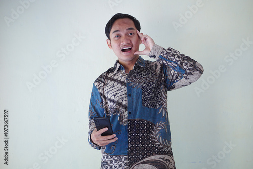 young Asian man wearing a batik shirt with a facial expression of surprise, surprise and amazement