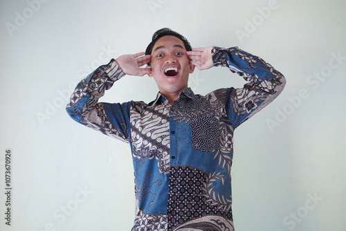young Asian man wearing a batik shirt with a facial expression of surprise, surprise and amazement