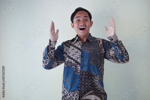 Excited young Asian man 20s wearing batik shirt pointing finger up, having great idea isolated on white background