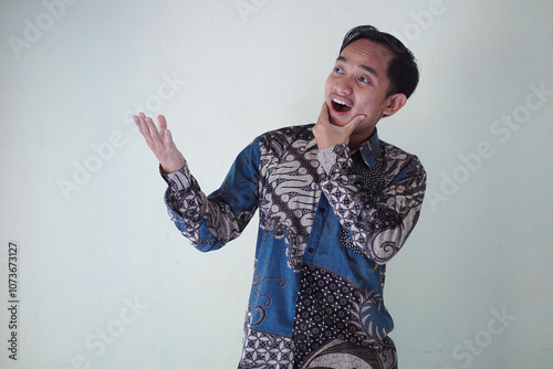 Excited young Asian man 20s wearing batik shirt pointing finger up, having great idea isolated on white background