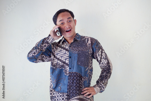 Thoughtful young asian man in a batik shirt holding a phone to his ear, looking puzzled and deep in thought against a plain background.