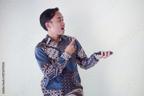 Shocked Asian man wearing batik shirt and looking at his cellphone screen, isolated on white background. expressing surprise and curiosity in a relaxed atmosphere.