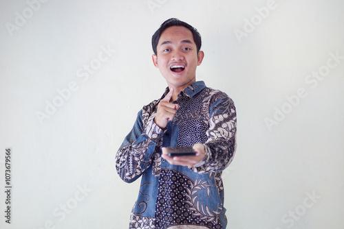 Shocked Asian man wearing batik shirt and looking at his cellphone screen, isolated on white background. expressing surprise and curiosity in a relaxed atmosphere.