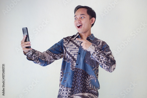 Shocked young Asian man wearing batik shirt looking at smartphone with surprised facial expression isolated on white background