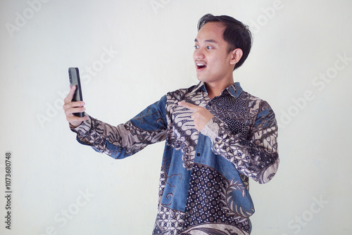 Shocked young Asian man wearing batik shirt looking at smartphone with surprised facial expression isolated on white background