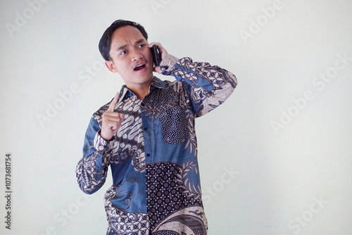 Thoughtful young asian man in a batik shirt holding a phone to his ear, looking puzzled and deep in thought against a plain background.