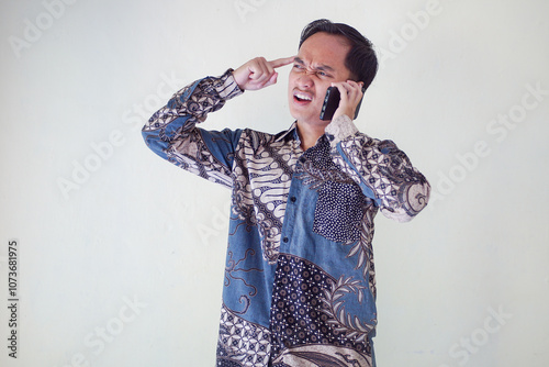 Thoughtful young asian man in a batik shirt holding a phone to his ear, looking puzzled and deep in thought against a plain background.