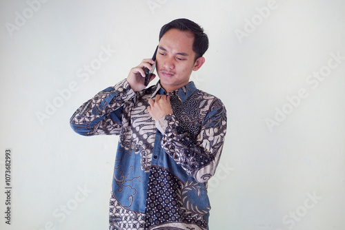 Thoughtful young asian man in a batik shirt holding a phone to his ear, looking puzzled and deep in thought against a plain background.