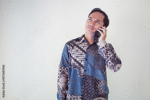 Thoughtful young asian man in a batik shirt holding a phone to his ear, looking puzzled and deep in thought against a plain background.