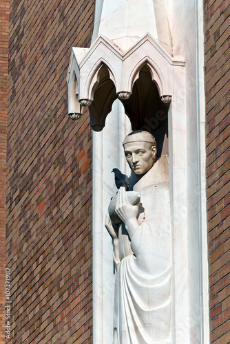 A statue on Szeged's Votive Church shows a monk holding a fish, with a live bird perched on it. The Gothic-inspired sculpture, set against a brick wall, captures a tranquil moment.