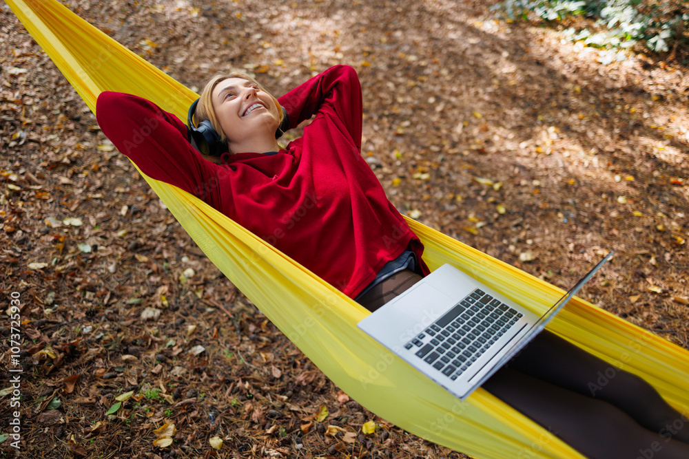Young woman in headphones with laptop relaxes while lying on a hammock in nature Young woman in headphones with laptop relaxes while lying on a hammock in nature