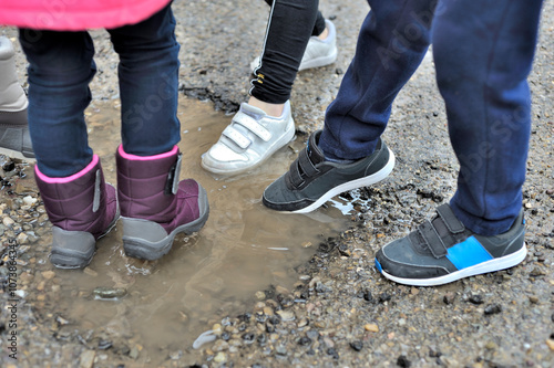 Horizontal photograph of 4 unrecognizable children sticking their feet in sports shirts and wellies in a small puddle of water. Selective focus. Copy space