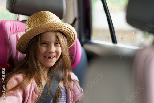 Little cute smiling girl without a front tooth in a sarafan and a straw hat sits fastened with a belt in a car. The concept of children's travel and recreation, health and dentistry photo