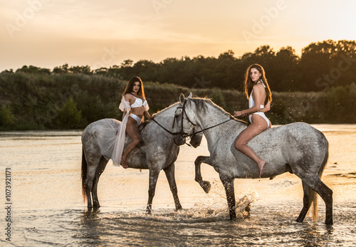 Obraz na płótnie Beautiful brunettes woman with long hair ,white bikini and white shirt rides on a white horse in the river. Sensual and attractive girls interacting and rides a horse in the water at sunset w salonie