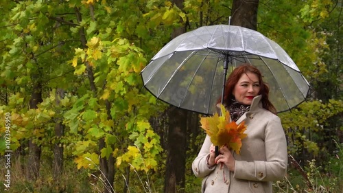 positive mature Caucasian woman stands with an umbrella and a bouquet of leaves in the park, smiling and having a great time.