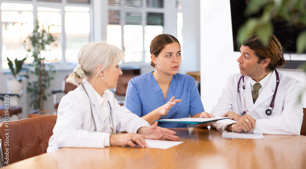 Focused confident young female doctor conducting medical council with experienced colleagues sitting at table with papers in office Focused confident young female doctor conducting medical council with experienced colleagues sitting at table with papers in office