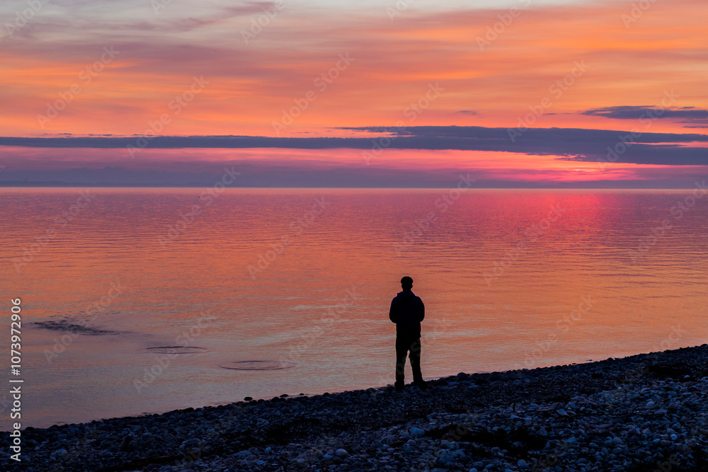 Fototapeta premium Silhouette of a man standing on a pebble beach at sunset
