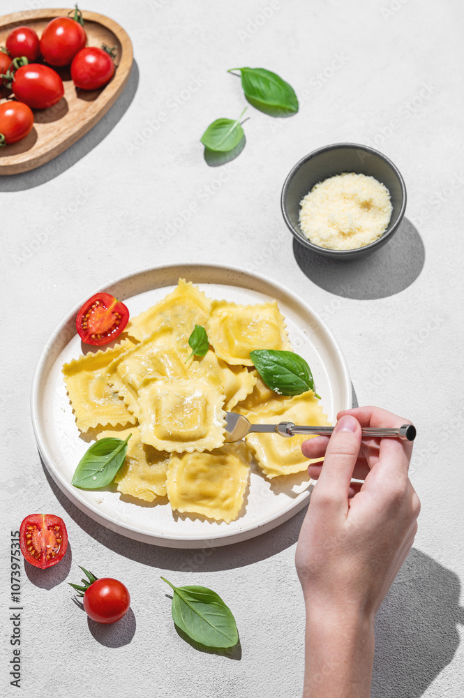 Hand holding fork in plate with homemade ravioli pasta with parmesan, tomatoes and basil on light background Hand holding fork in plate with homemade ravioli pasta with parmesan, tomatoes and basil on light background