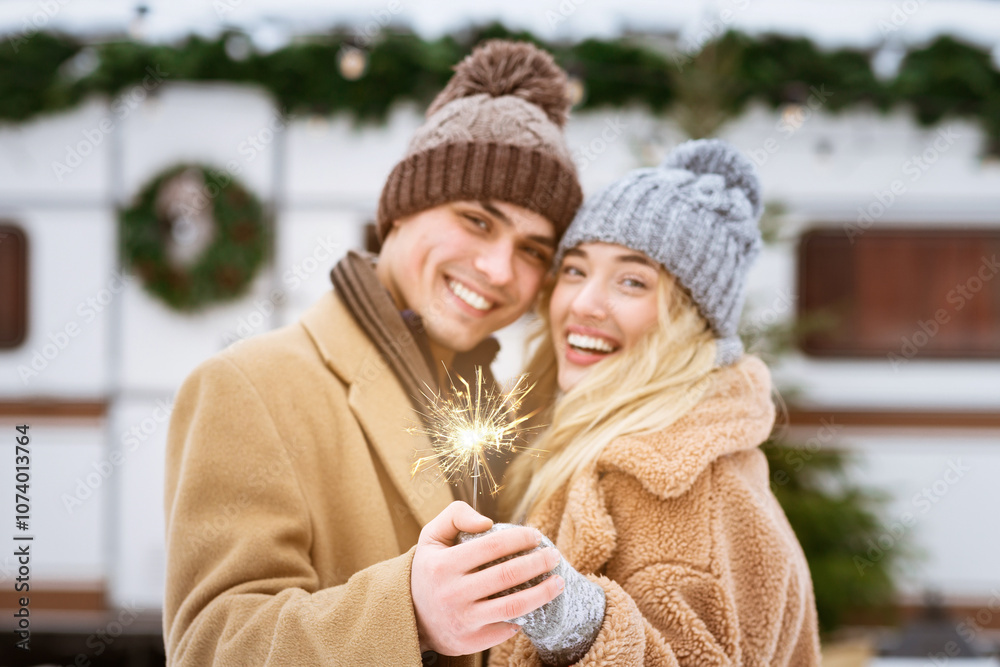A young couple smiles joyfully while holding a sparkling sparkler. They are dressed warmly for winter and surrounded by festive decorations outdoors. A young couple smiles joyfully while holding a sparkling sparkler. They are dressed warmly for winter and surrounded by festive decorations outdoors.