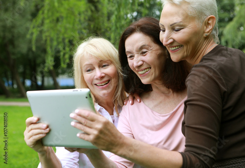 Three old women are smiling and looking photos at the at screen of the tablet pc in the park on a summer day. Friendship is priceless.