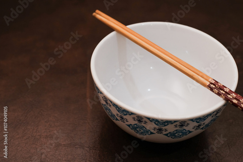 White bowl with blue pattern outside and wooden chopsticks on brown background, traditional wooden sticks for asian food.