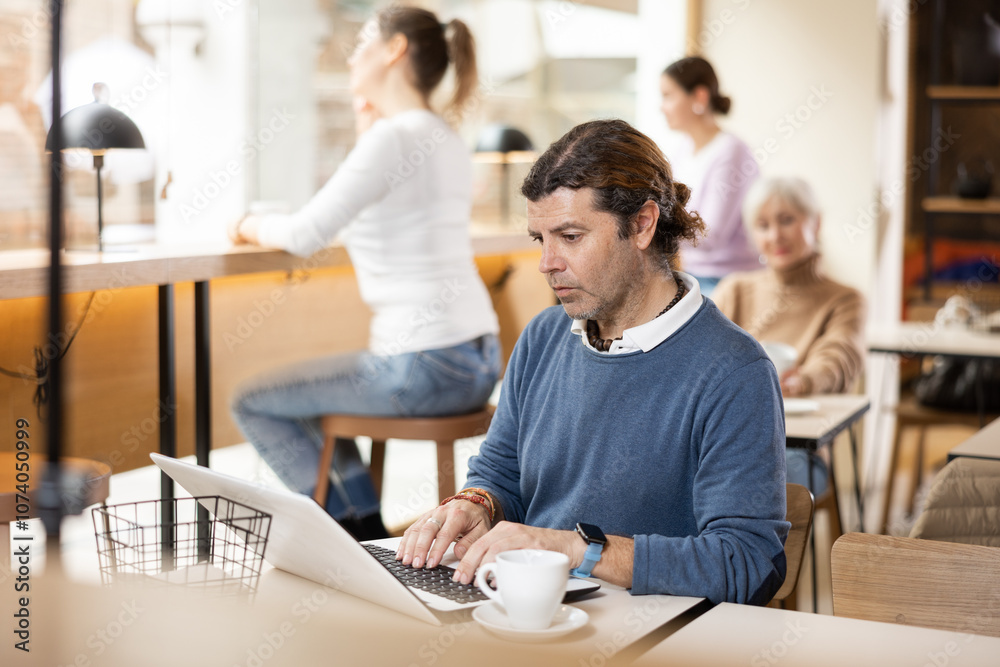 Serious middle-aged man sitting at table, using laptop and enjoing cup of tea in coffee house Serious middle-aged man sitting at table, using laptop and enjoing cup of tea in coffee house