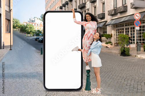 Two friends are having fun in a vibrant city, one standing on a pole and the other next to her, beside a large blank billboard. The surroundings include charming buildings and greenery. photo