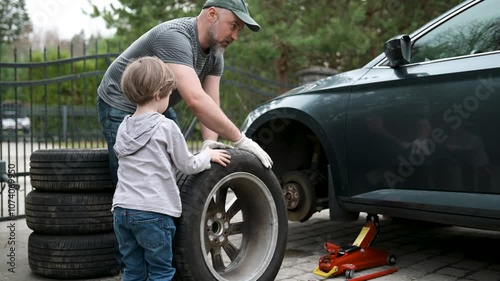 Cute toddler boy helping his father to change car wheels in their backyard. Father teaching his little son motor vehicle construction basics. Small child active parent. Slow motion summer day footage.