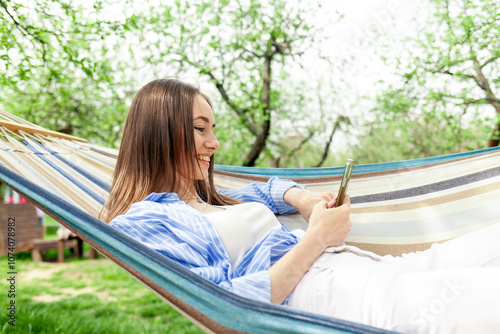 young cheerful girl lies on comfortable hammock in garden in summer and uses smartphone online, woman relaxes in park outdoors and looks at mobile phone and smiles