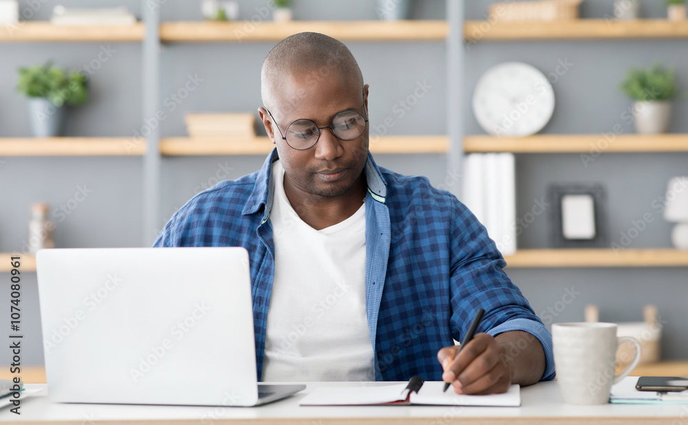 Concentrated black mature man attending online training from home in front of laptop and taking notes, sitting in home office, copy space. E-education, online class concept Concentrated black mature man attending online training from home in front of laptop and taking notes, sitting in home office, copy space. E-education, online class concept