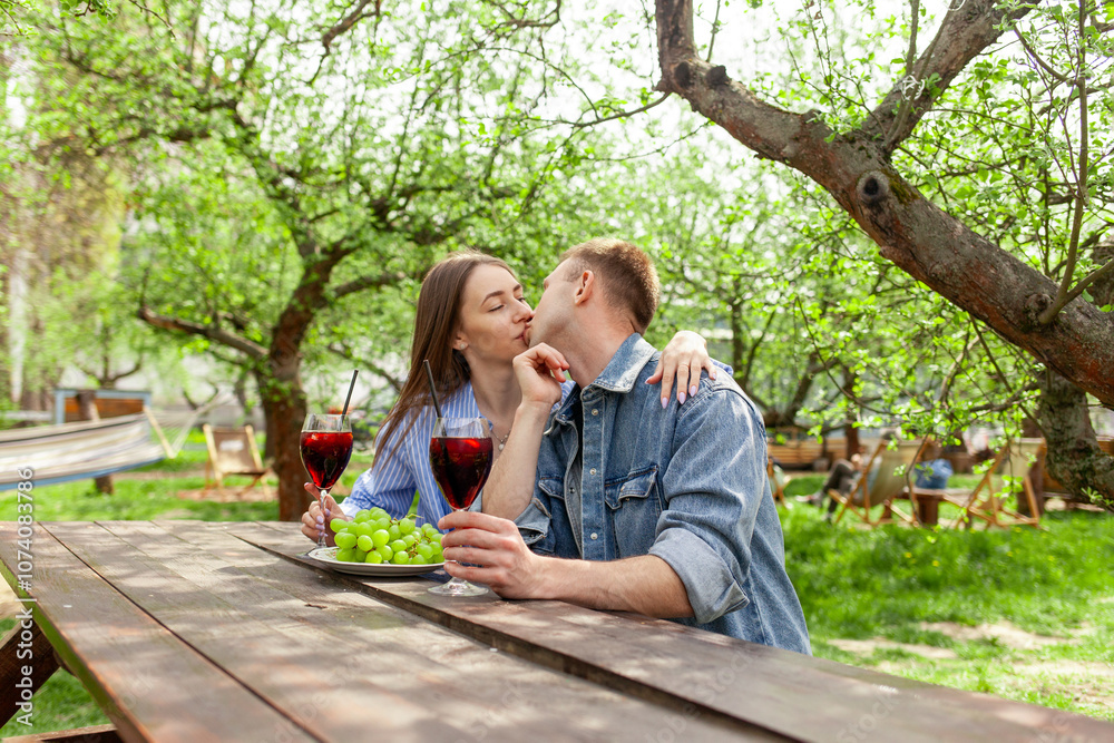 Naklejka premium young couple in love in denim clothes sitting in the garden at wooden table and drinking red wine with grapes on date and kissing in the summer, guy and girl trying alcoholic cocktails and flirting in