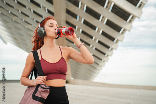 Young athletic woman in sports wear and headphones recovering drinking isotonic water from bottle after outdoor cardio workout training session. Female hydratating after her wellbeing gym rutine photo