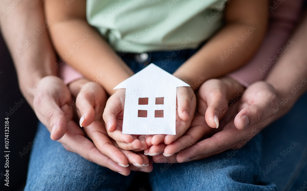 Man, woman and little child holding cutout paper house figure in hands, conceptual image for family housing, home mortgage, real estate, insurance or adoption, closeup shot Man, woman and little child holding cutout paper house figure in hands, conceptual image for family housing, home mortgage, real estate, insurance or adoption, closeup shot
