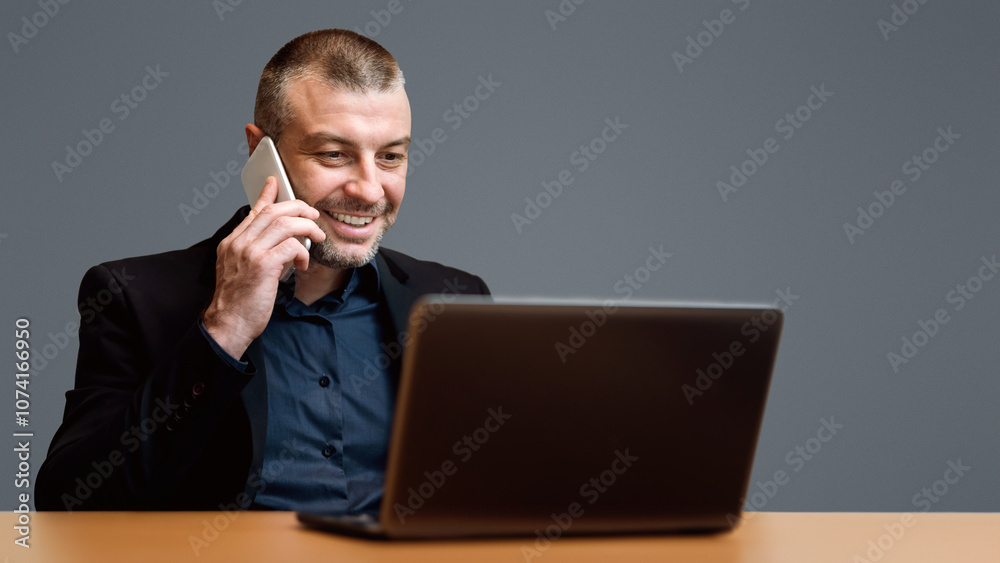 Businessman Working On Laptop Talking On Cellphone Sitting Over Gray Background. Studio Shot, Selective Focus Businessman Working On Laptop Talking On Cellphone Sitting Over Gray Background. Studio Shot, Selective Focus