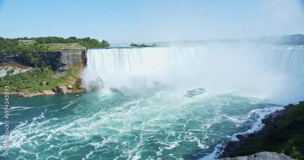 A breathtaking view of Niagara Falls on a sunny summer day, with mist rising as the water cascades down. In the distance, a tour boat approaches the powerful waterfall. A handheld shot. A breathtaking view of Niagara Falls on a sunny summer day, with mist rising as the water cascades down. In the distance, a tour boat approaches the powerful waterfall. A handheld shot.