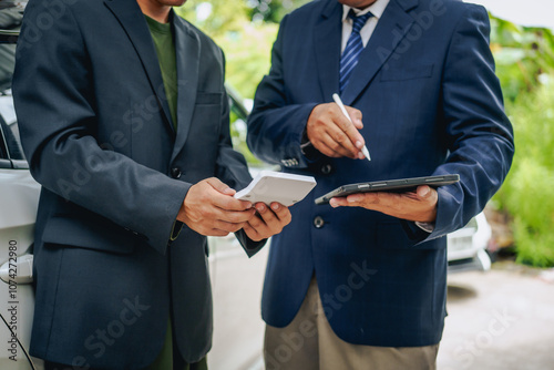 Asian clients engage with a car insurance agent beside a wrecked vehicle. They examine the policy details, assess the damage, and finalize paperwork related to the insurance claim and repairs.