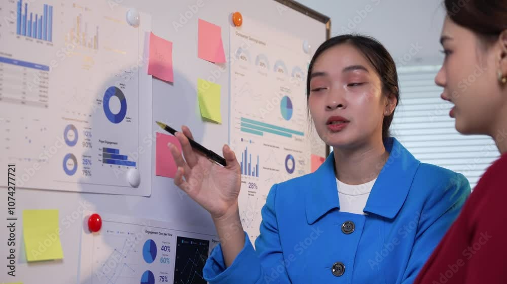 Businesswoman in a blue blazer stands at a whiteboard presenting data analysis to a colleague in a red blazer Businesswoman in a blue blazer stands at a whiteboard presenting data analysis to a colleague in a red blazer