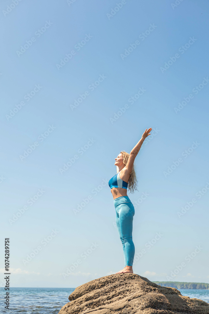 Obraz premium Smiling fitness yoga woman standing on cliff over sea raising hands enjoy healthy lifestyle at beach